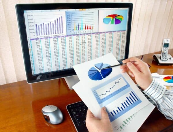 A person holds up documents containing graphs and checks them against their computer which displays financial figures.