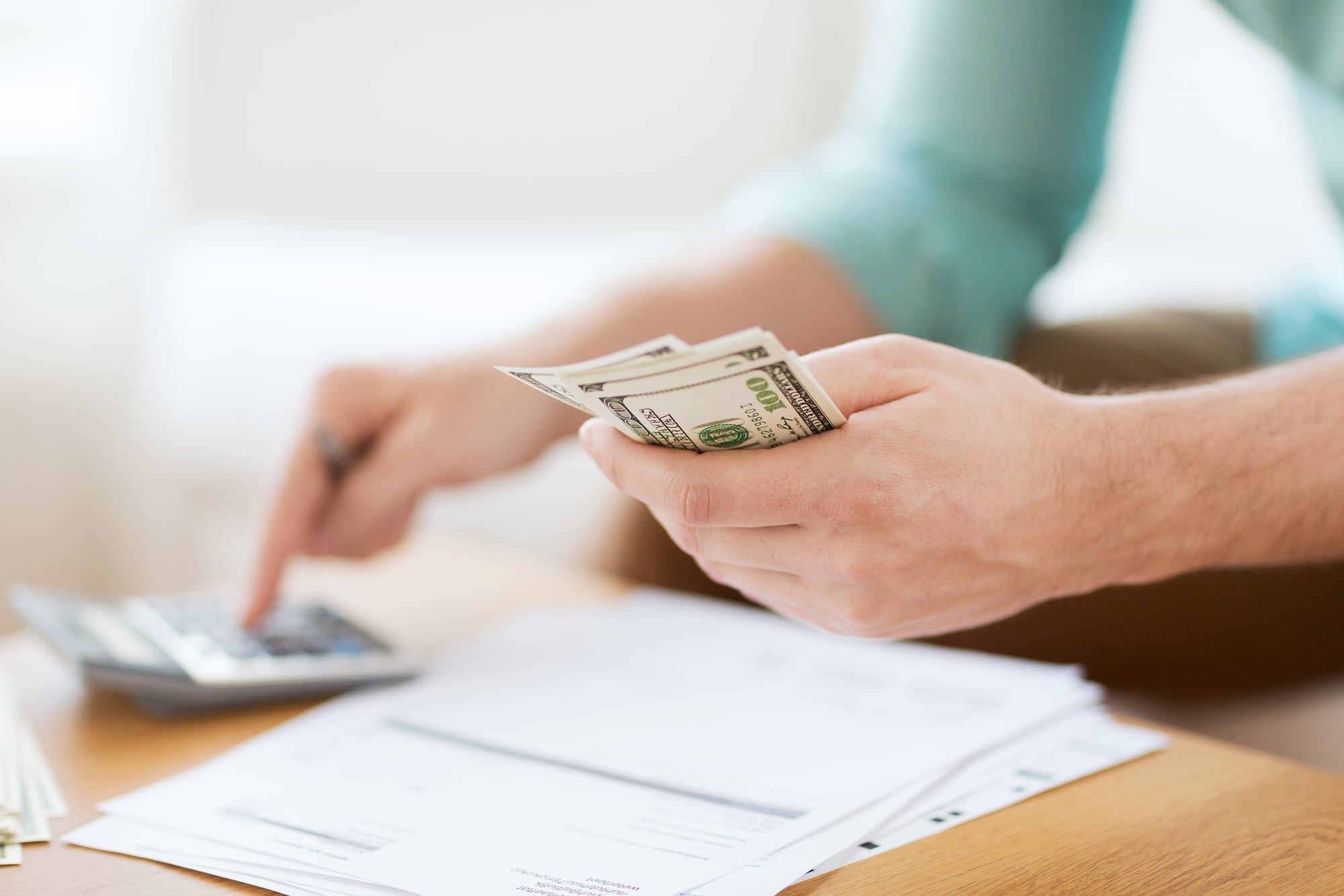 A closeup of a person sitting at a desk with financial documents in front of them, typing on a calculator with one hand while holding money in the other.