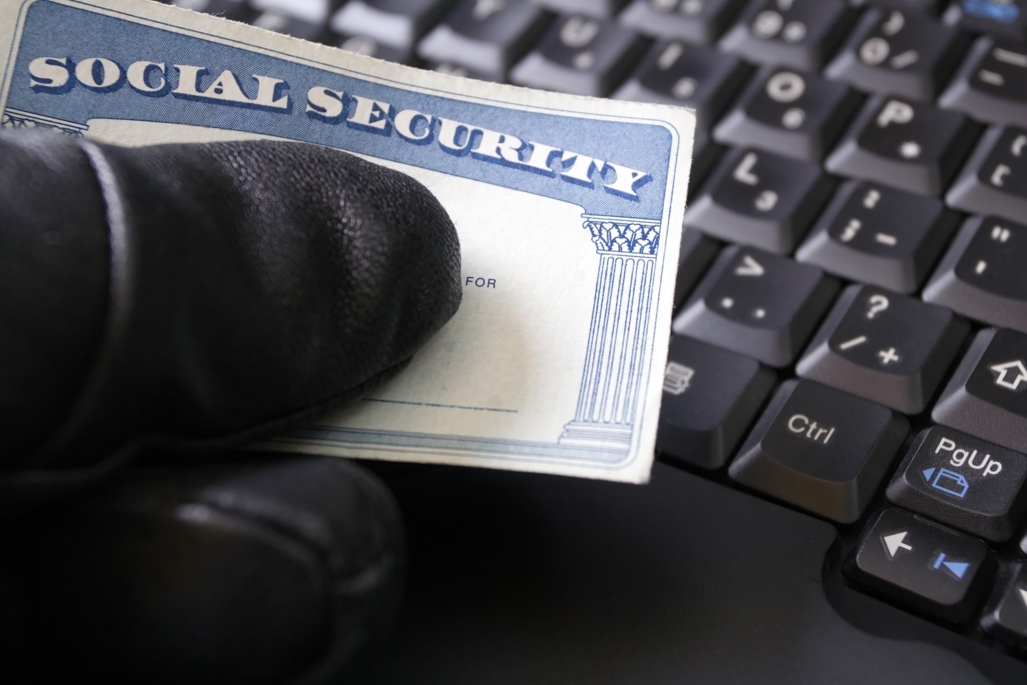 Extreme close-up of a black-gloved hand holding a Social Security card above a computer keyboard.